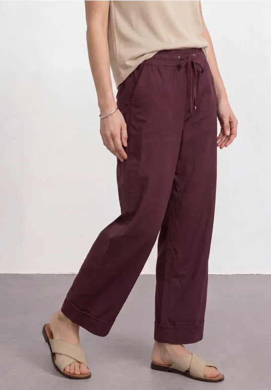 Weite burgunderrote Leinenhose mit Tunnelzug, seitlichen Taschen, kombiniert mit hellem Top und bequemen Sandalen.