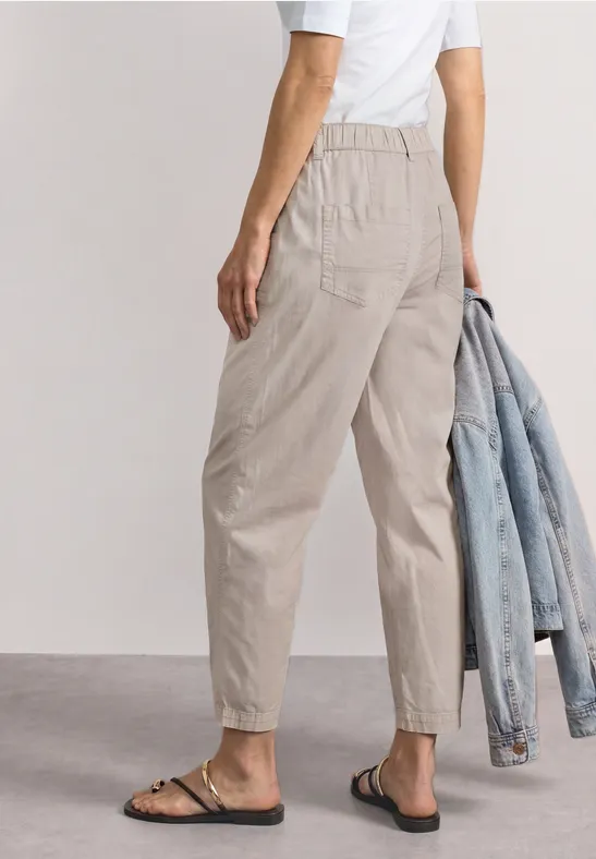 Person von hinten in beigen Bundfaltenhosen, weißem Shirt und Sandalen, hält Jeansjacke.