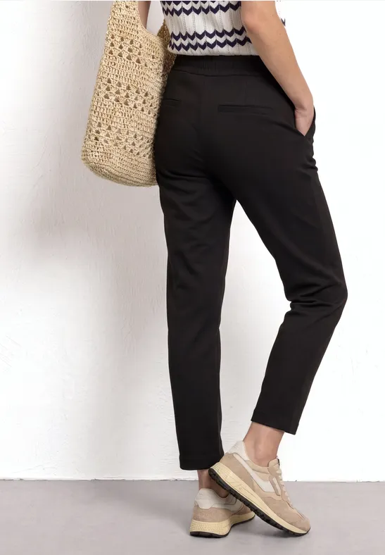 Vrouw van achteren in zwarte chino, gestreepte top, rieten tas en beige sneakers.