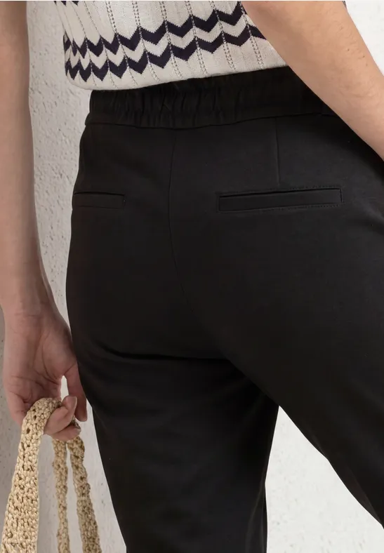 Detail van iemand in zwarte broek met elastische taille en achterzak, die een gehaakte tas vasthoudt.
