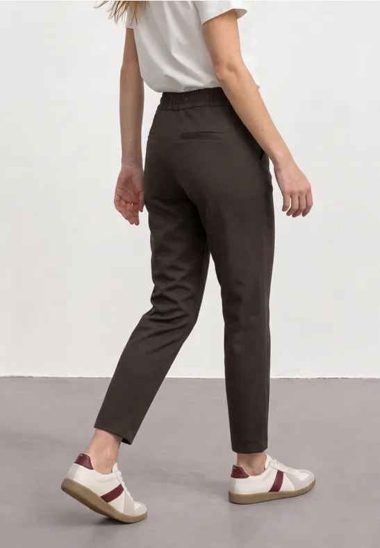 Frau trägt schmale, dunkelbraune Hose und weiße Sneaker mit rotem V-Design vor heller Wand.