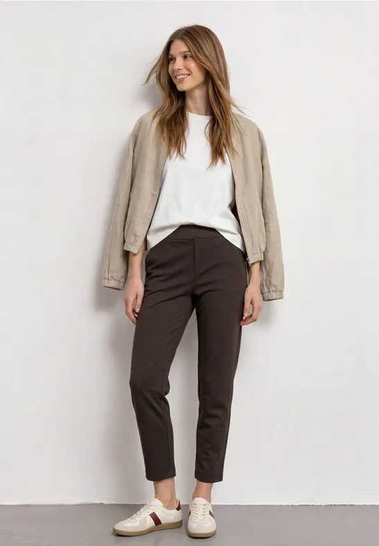 Eine junge Frau in weißem Shirt, beigem Blazer, dunklen Hosen und Sneakers vor hellgrauem Hintergrund.
