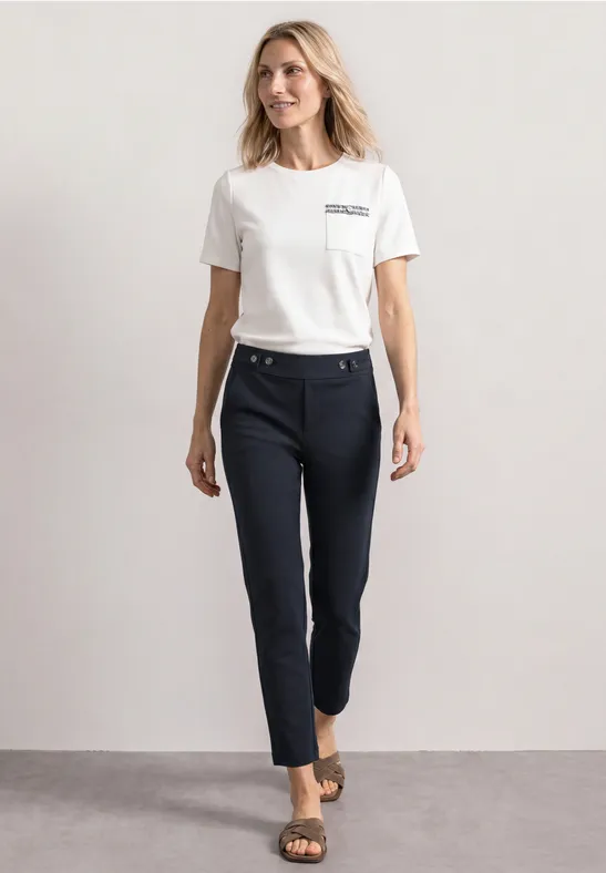 Frau mit weißem Shirt, dunkelblauen knöchellangen Hose und Sandalen vor neutralem Hintergrund