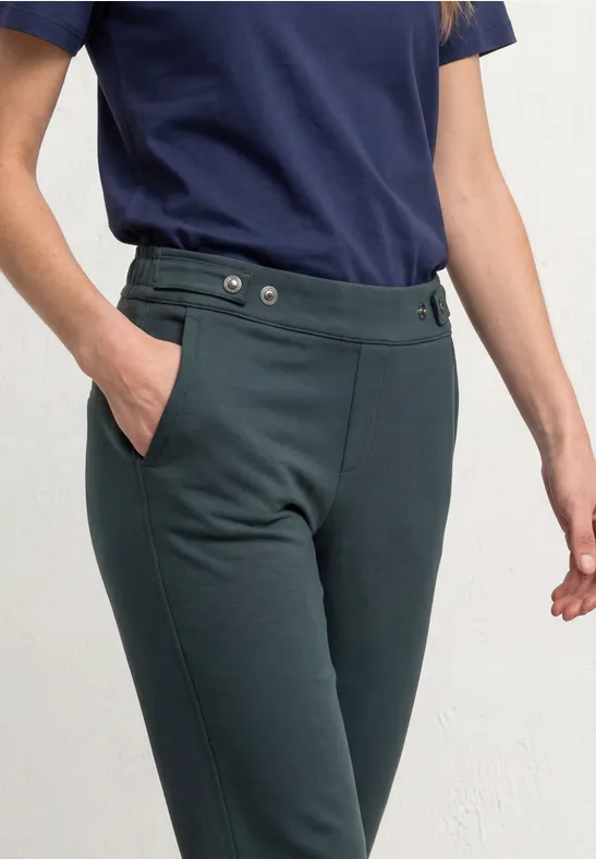 Vrouw draagt een donkergroene, getailleerde broek met knopen en zijzakken, gecombineerd met een marineblauw t-shirt.