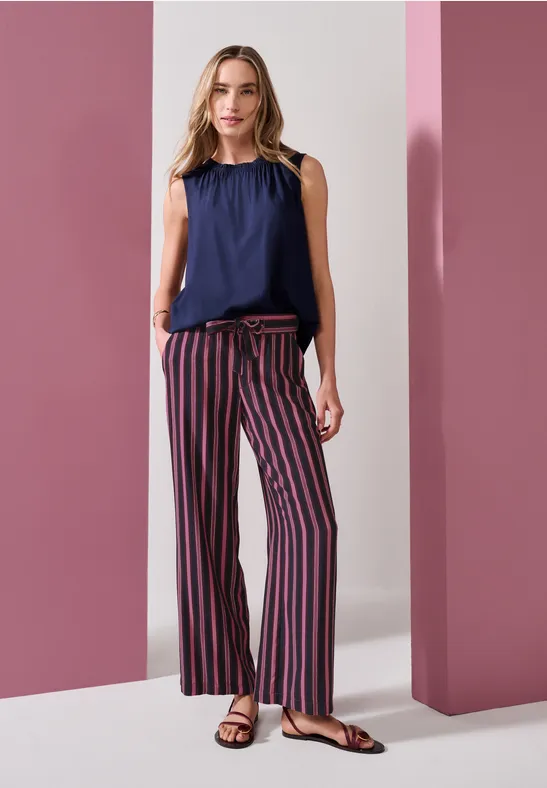 Frau in navy-blauer ärmelloser Bluse und gestreifter Marlene-Hose vor rosa Säulen.