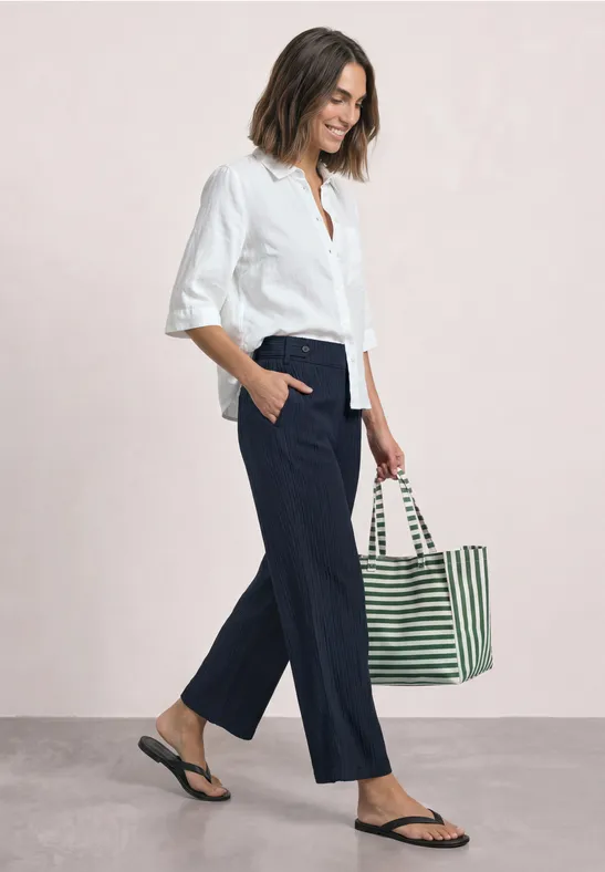 Eine Frau in weißem Hemd, blauer Culotte, Flipflops und gestreifter Tasche, im Profil gehend.