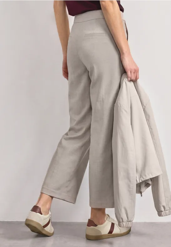 Model draagt wijde grijze broek, houdt bijpassend colbert vast en combineert dit met plateau-sneakers.
