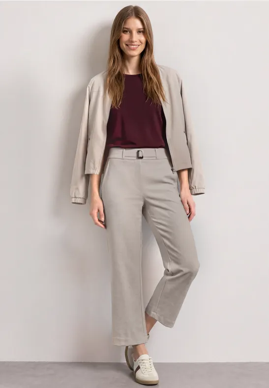 Vrouw in beige cropped broek met bijpassend jasje over de schouders, bordeaux top en witte sneakers.
