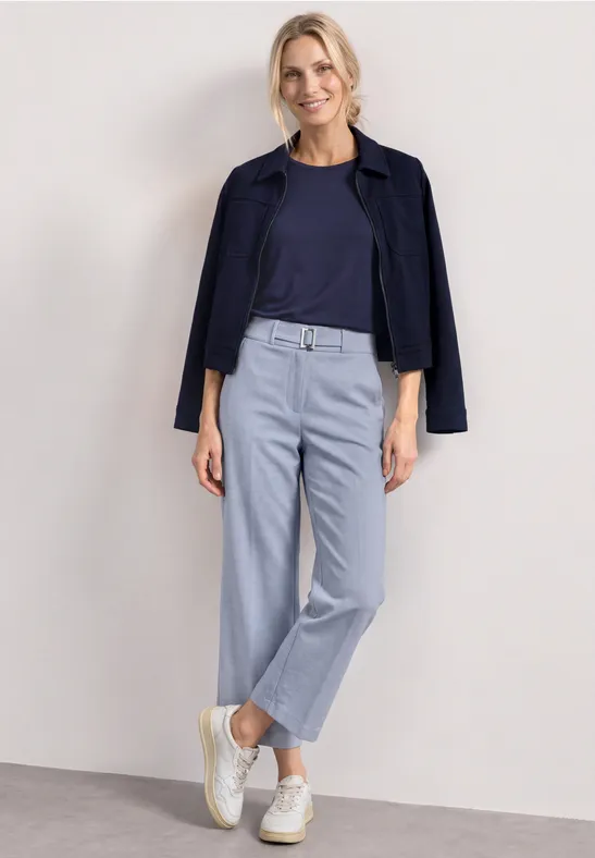 Een vrouw in een marineblauw topje en jasje, lichtblauwe broek en witte sneakers tegen een neutrale achtergrond.