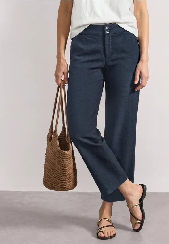Vrouw met wit hemd, wijde donkerblauwe broek, gevlochten tas en sandalen.