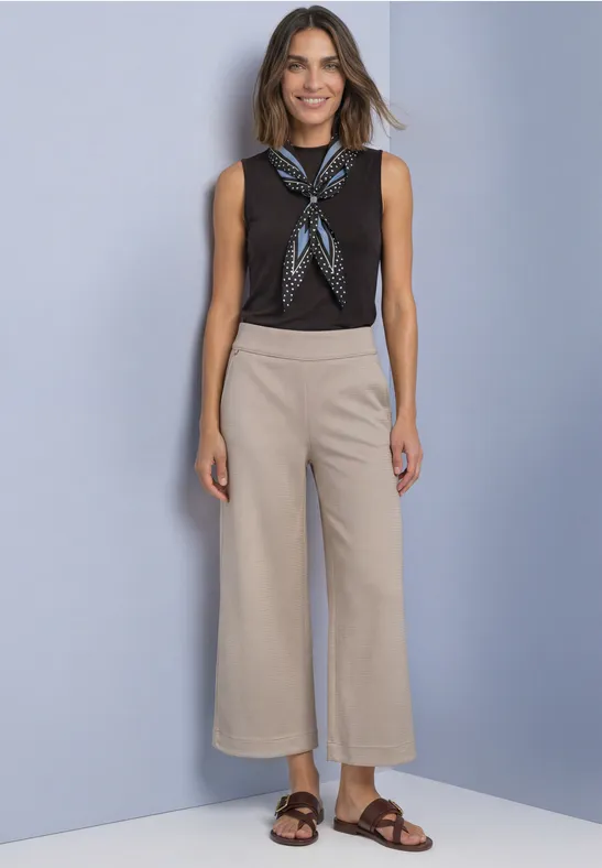 Frau in schwarzem Top, gemustertem Halstuch, beiger Culotte und braunen Sandalen vor blauem Hintergrund.