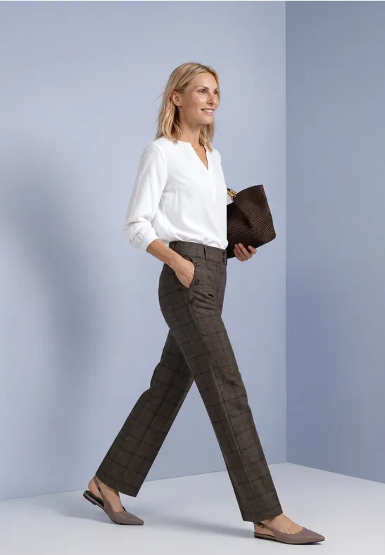 Frau in weißer Bluse und braunen Karohosen, geht mit einer braunen Clutch in der Hand.