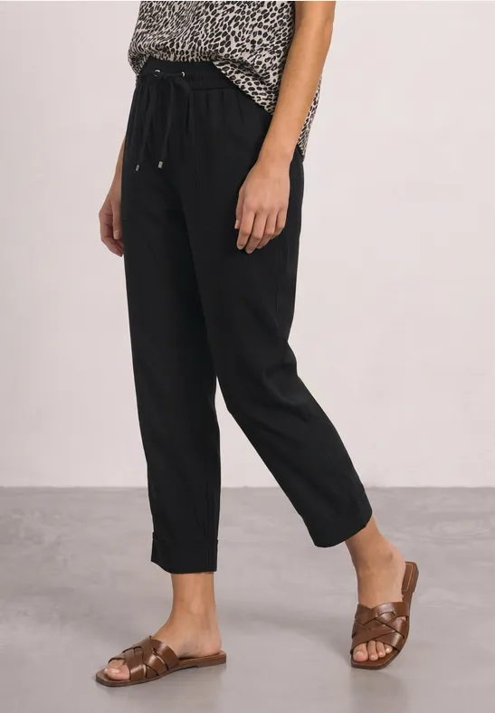 Vrouw draagt zwarte, enkellange broek met losse pasvorm en bruine, platte sandalen.