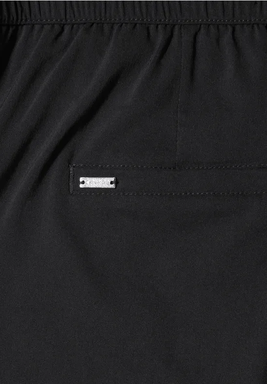 Close-up van een zwarte broek met elastische tailleband en een klein metalen plaatje bij de zak.