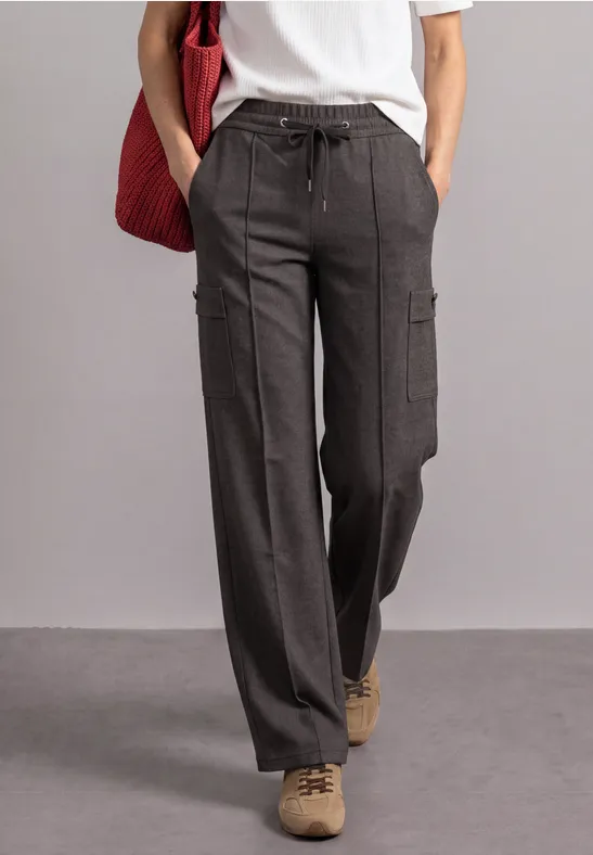 Person trägt weite graue Cargo-Hose, weißes T-Shirt, beigen Schuhe, hält rote Tasche.