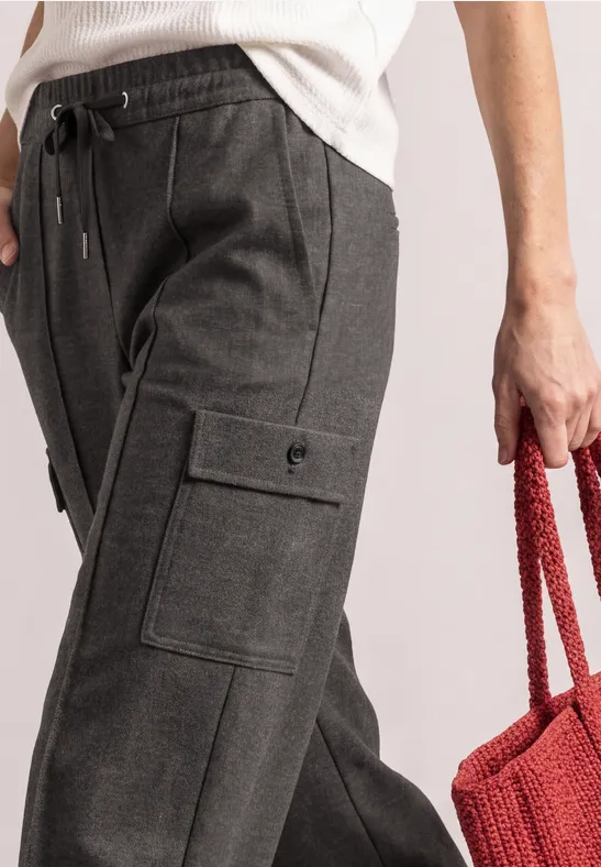 Dunkelgraue Cargohose mit seitlichen Pattentaschen und rotem Strickbeutel in der Hand.