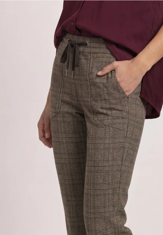 Eine Person trägt karierte, hoch taillierte Stoffhose mit Kordelzug und Eingrifftaschen in Braun.