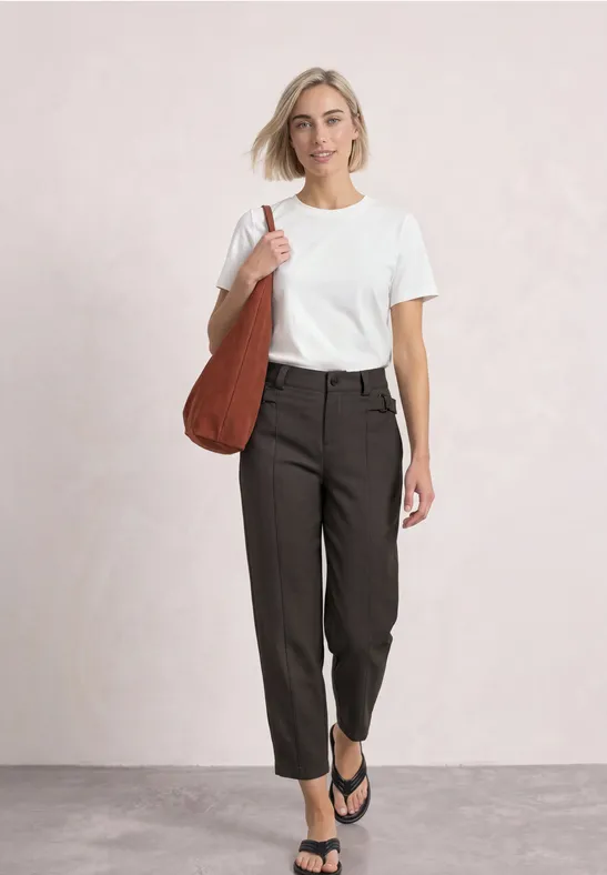 Frau im weißen T-Shirt, dunkler Hose und Flip-Flops mit brauner Schultertasche.