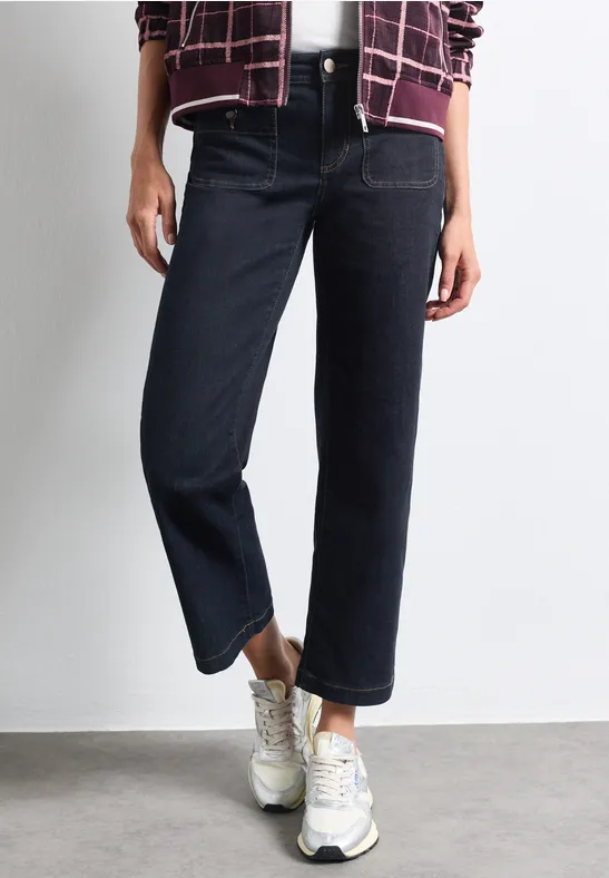 Person trägt dunkle, gerade geschnittene Jeans und weiße Sneakers, stehende Pose auf grauem Untergrund.