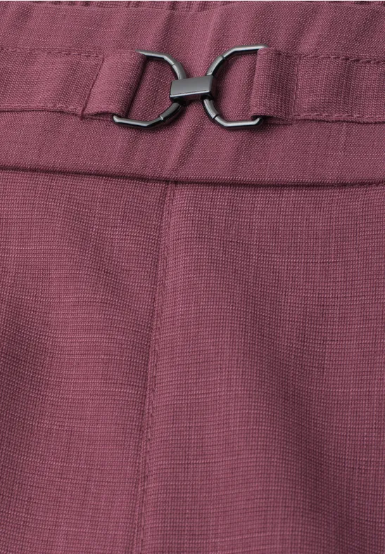 Gros plan d’un pantalon couleur prunelle à la texture délicate, agrémenté d’une boucle métallique argentée.