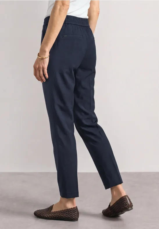 Person trägt dunkelblaue, gerade geschnittene Hose mit elastischem Bund und geflochtenen Loafers.