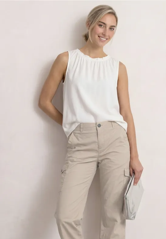 Junge Frau in beige Hose und weißem ärmellosen Oberteil, hält eine hellgraue Clutch.