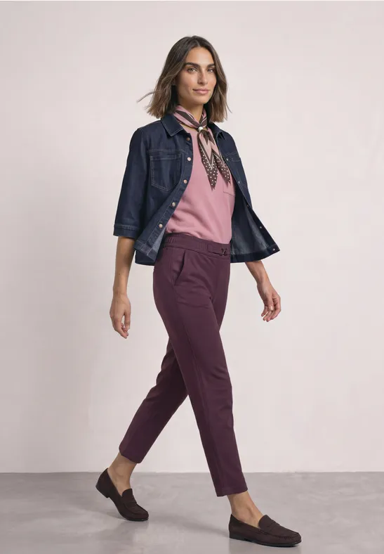 Frau im stylischen Outfit: rosa Bluse, dunkelrote Hose, Jeansjacke, Halstuch und schwarze Loafers.