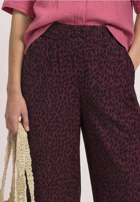 Frau trägt weinrote Hose mit schwarzem Leoprint, rosafarbenes Oberteil und hält eine gehäkelte Tasche.