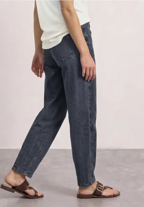 Eine Person trägt dunkle, lockere Jeans und braune Sandalen, seitliche Ansicht.