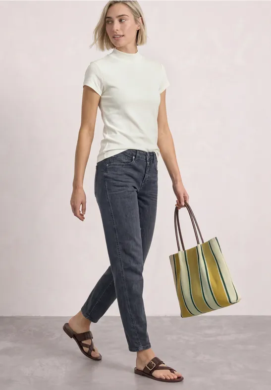 Frau in weißem T-Shirt, dunkler Jeans, Sandalen und gestreifter Tragetasche vor hellem Hintergrund.