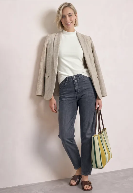 Frau in weißem Pullover, beigem Blazer, dunklen Jeans, Sandalen und gestreifter Tasche vor neutralem Hintergrund.