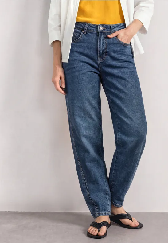 Eine Person trägt blaue Jeans, gelbes Oberteil, weiße Jacke und schwarze Sandalen.