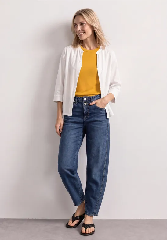 Frau in weißer Jacke, gelbem Shirt, blauen Jeans und schwarzen Sandalen vor neutralem Hintergrund.