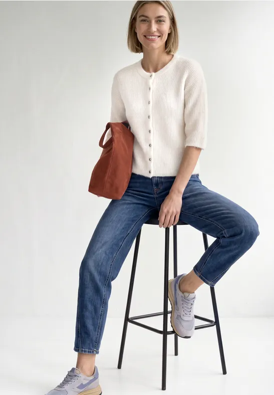 Frau sitzt auf Hocker, trägt cremefarbenen Cardigan, blaue Jeans, weiße Sneaker und hält eine braune Tasche.