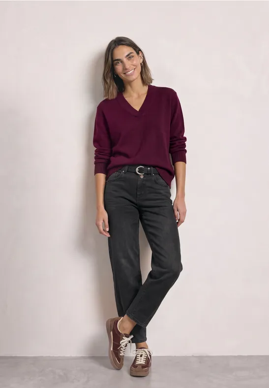 Vrouw in paarse trui, zwarte jeans en bruine schoenen poseert voor een lichte muur.