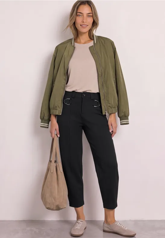 Frau trägt grüne Bomberjacke, helles Top, schwarze Hose und große Beuteltasche.