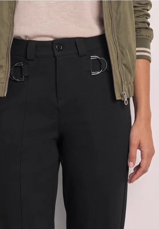 Schwarze Hose mit D-Ringen an der Taille, kombiniert mit beigem Shirt und grüner Jacke.