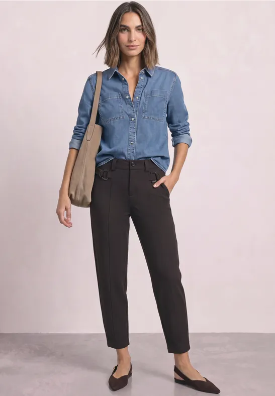 Eine Frau in Jeanshemd, schwarzen Hosen und Slingbacks trägt eine Schultertasche vor hellem Hintergrund.