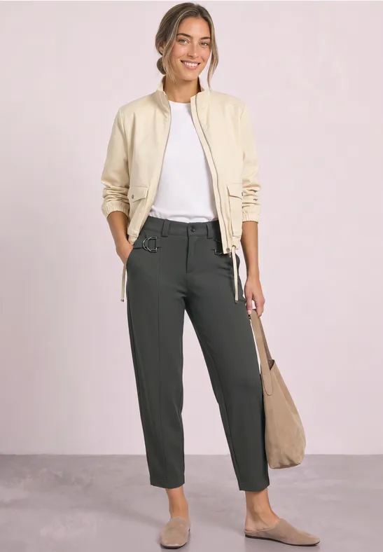 Frau mit heller Jacke, weißem Shirt und dunklen Hosen trägt Beutetasche und lächelt vor hellem Hintergrund.