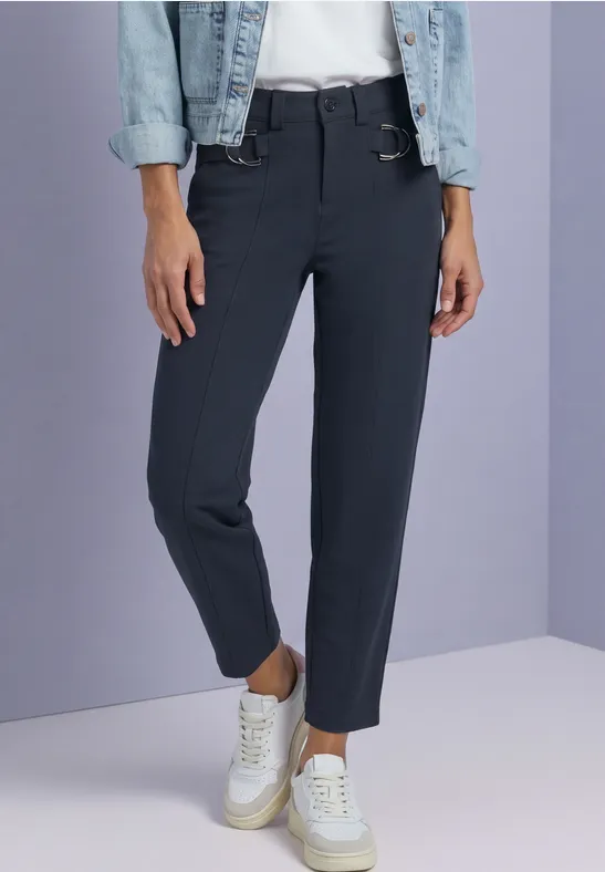 Schwarze 7/8-Hose und weiße Sneaker, kombiniert mit einer Jeansjacke vor fliederfarbenem Hintergrund.