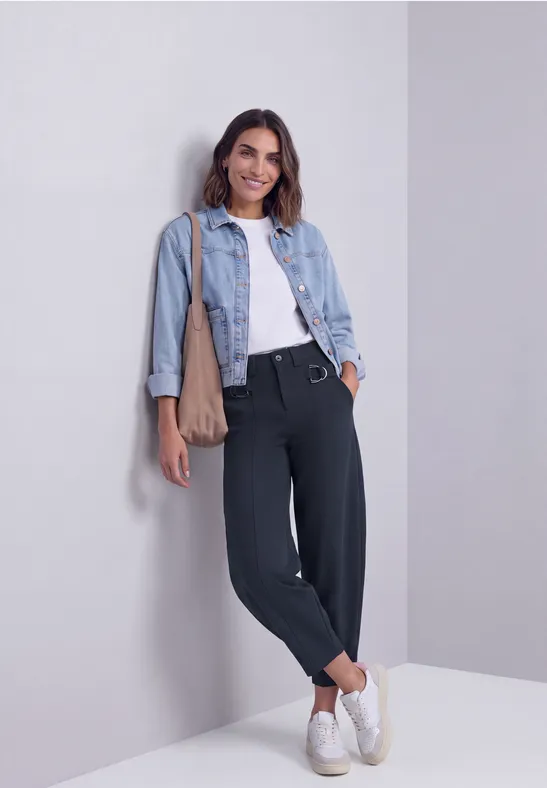 Junge Frau lehnt lässig an einer Wand, trägt Jeansjacke, weißes Top, schwarze Hose und weiße Sneaker.