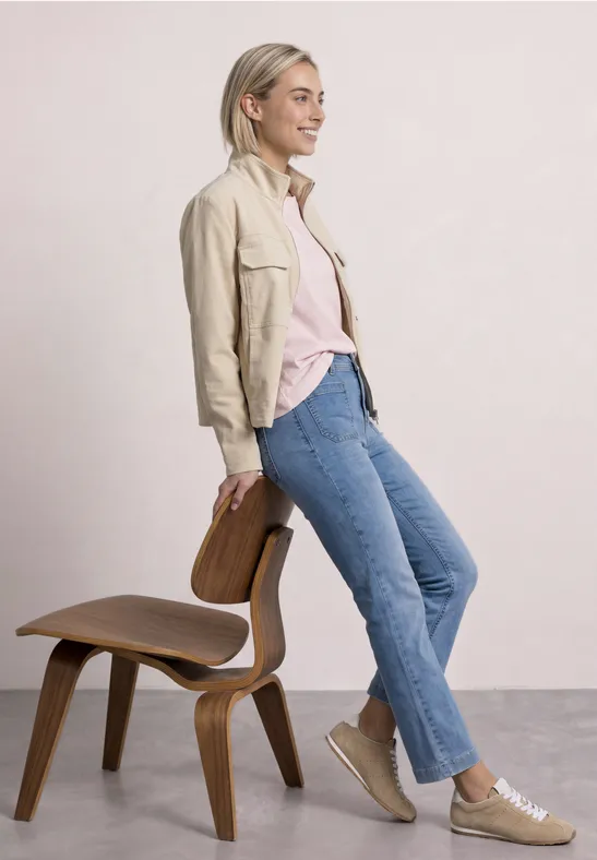 Een vrouw leunt nonchalant tegen een houten stoel, draagt een lichte jas, spijkerbroek en sneakers.