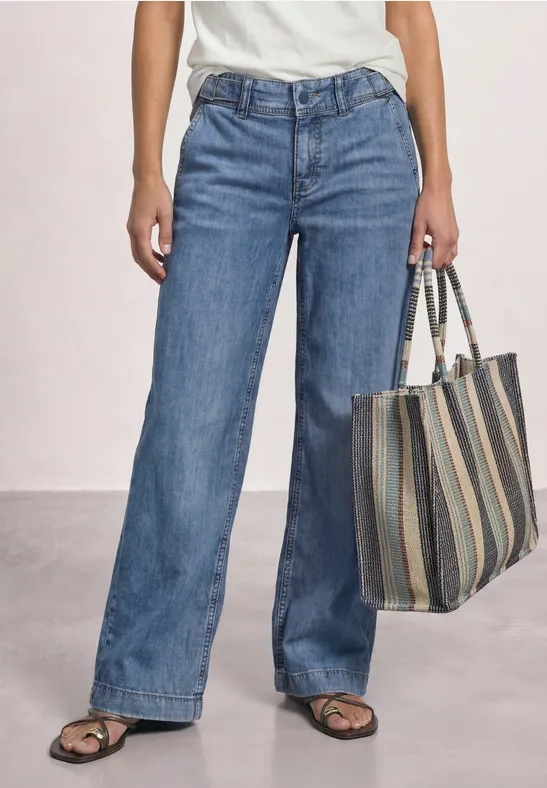 Vrouw in wijde spijkerbroek, wit topje en sandalen draagt gestreepte tas.