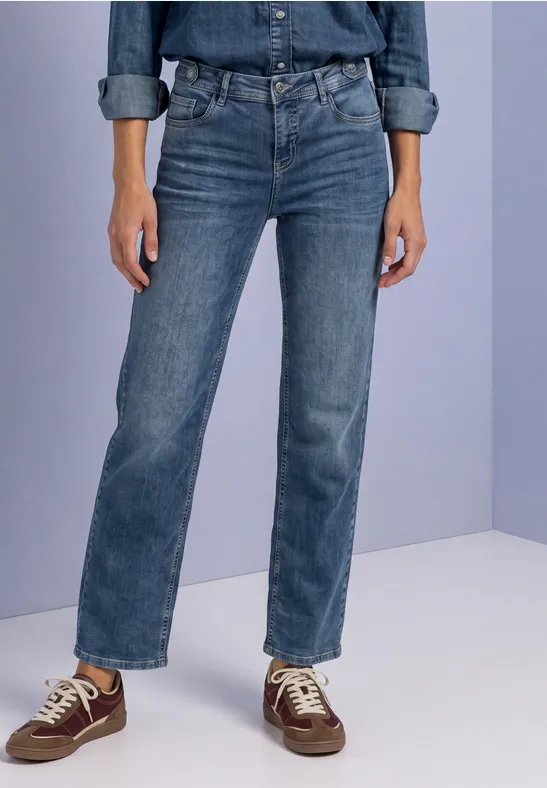 Eine Person steht vor hellem Hintergrund, trägt blaue Jeans und braune Sneaker.
