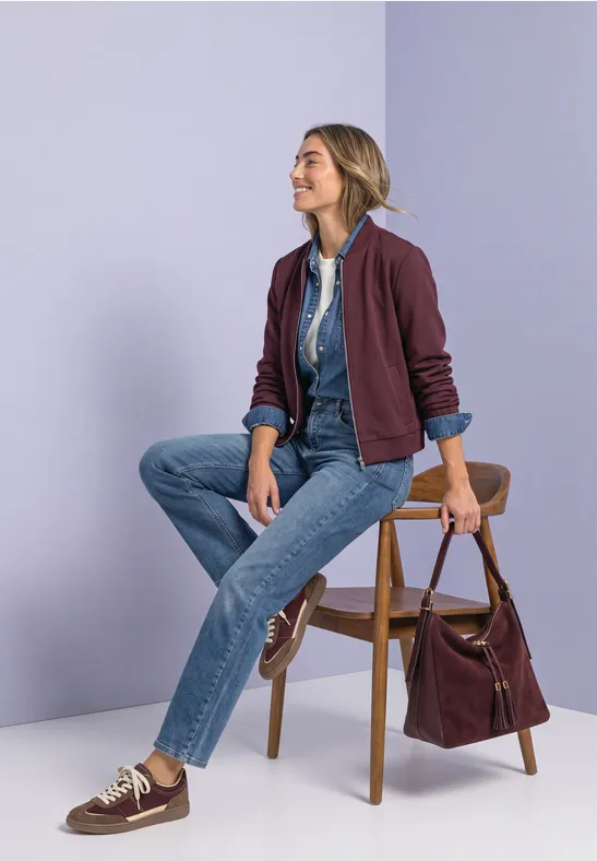 Eine Frau sitzt entspannt auf einem Stuhl, trägt Jeans, Hemd und bordeauxrote Jacke, daneben eine Tasche.