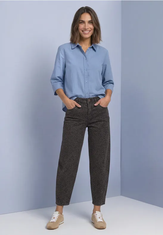 Eine lächelnde Frau in hellblauer Bluse und braunen, gemusterten Hosen mit Sneakers vor hellblauem Hintergrund.