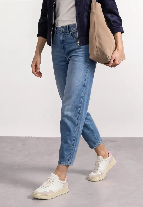 Persoon in blauwe jeans, witte sneakers en een donkerblauwe jas draagt een katoenen tas over de schouder.