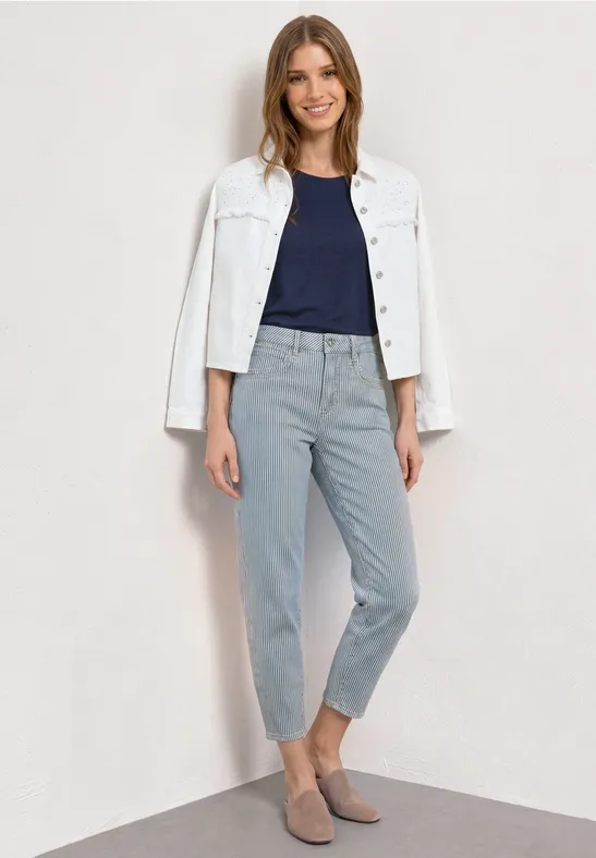 Femme en veste blanche sur t-shirt bleu marine et pantalon gris clair, debout devant un mur clair.