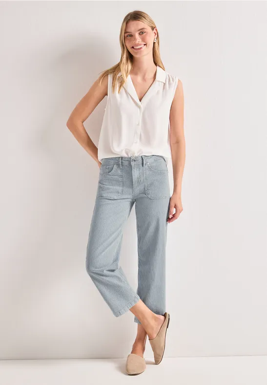 Femme en chemisier blanc sans manches, jean bleu clair et mocassins beiges sur fond clair.