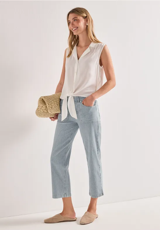 Femme en chemise blanche nouée, jean bleu clair, mocassins beiges et sac en paille devant un mur blanc.